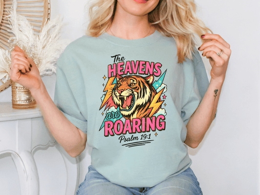 A woman wearing a light blue t-shirt with a graphic design of a tiger and the text "The Heavens are Roaring" printed on it.