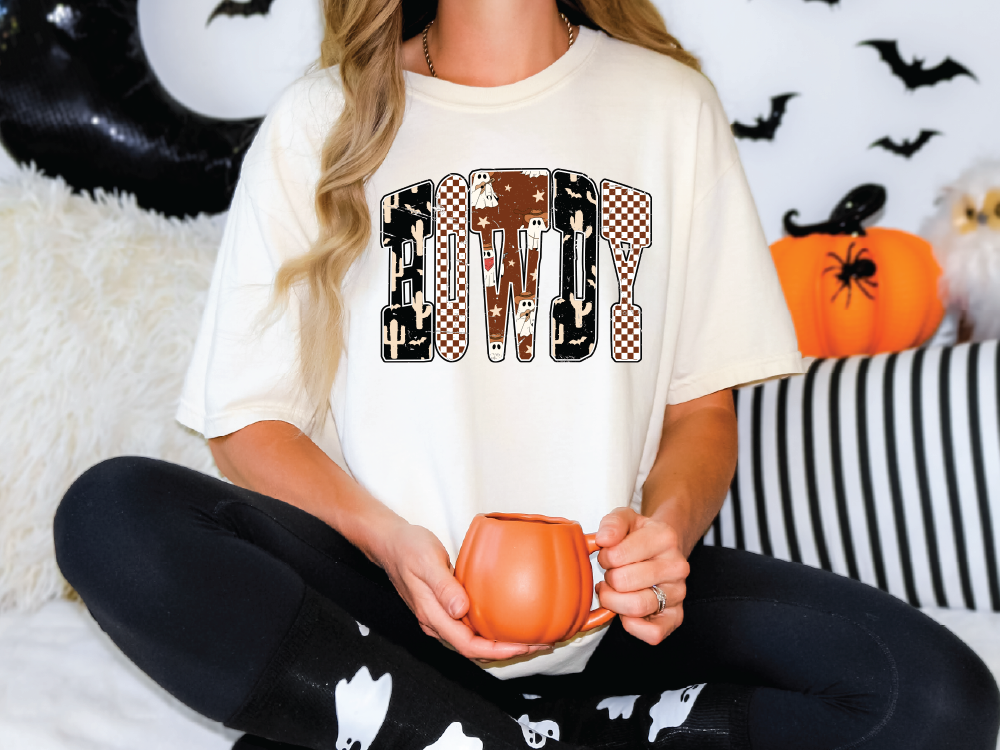 A woman wearing a white t-shirt with the word "BOWDY" printed on it, sitting on a couch with a pumpkin-shaped mug in her hands.