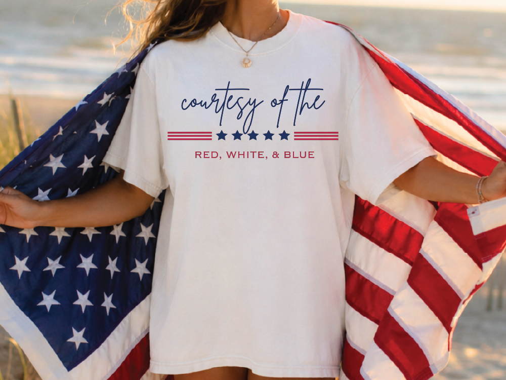 A person wearing a white t-shirt with the text "courtesy of the" printed on it, standing on a beach with an American flag draped over their shoulder.