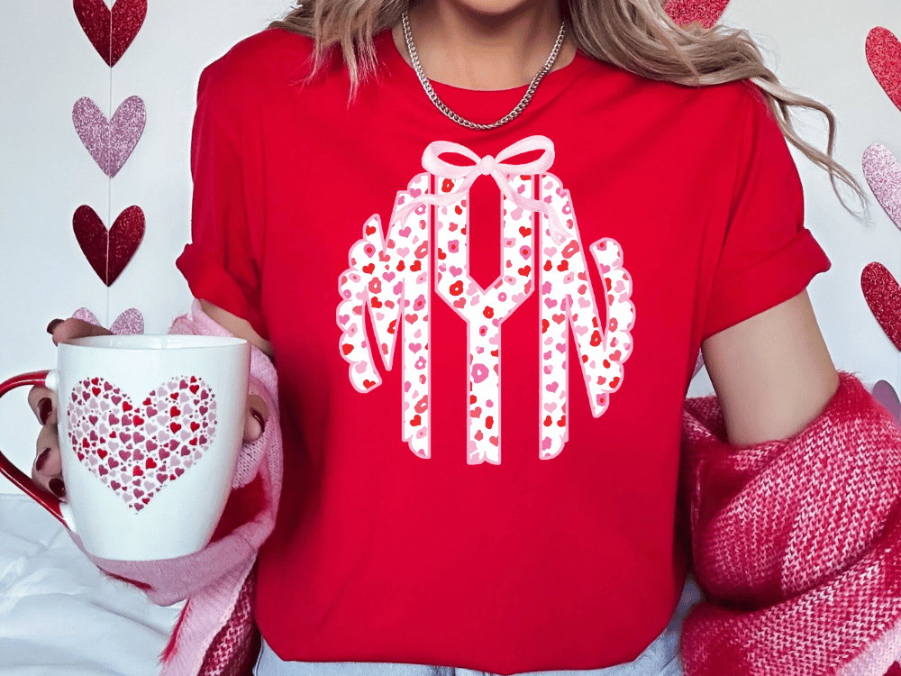 A woman wearing a red t-shirt with a monogram design and holding a white mug with a heart pattern.