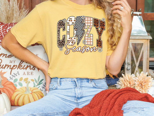 A person wearing a yellow t-shirt with the word "COZY" printed on it, sitting on a couch with a red blanket and a pumpkin decoration.