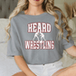 A woman wearing a gray t-shirt with the text "HEARD WRESTLING" printed on it, sitting on a bed.