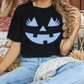 A woman wearing a black t-shirt with a blue jack-o-lantern design is sitting on a couch surrounded by fall decorations.