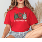 A woman wearing a red t-shirt with a Christmas tree design and the text "LET HEAVEN NATURE SING" is sitting with her legs crossed.