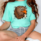 A person wearing a turquoise t-shirt with a graphic of a brown eagle and a star on it, sitting on the floor.
