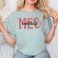 A woman wearing a light blue t-shirt with the text "MES" printed on it, sitting in front of a white wall with a decorative item behind her.
