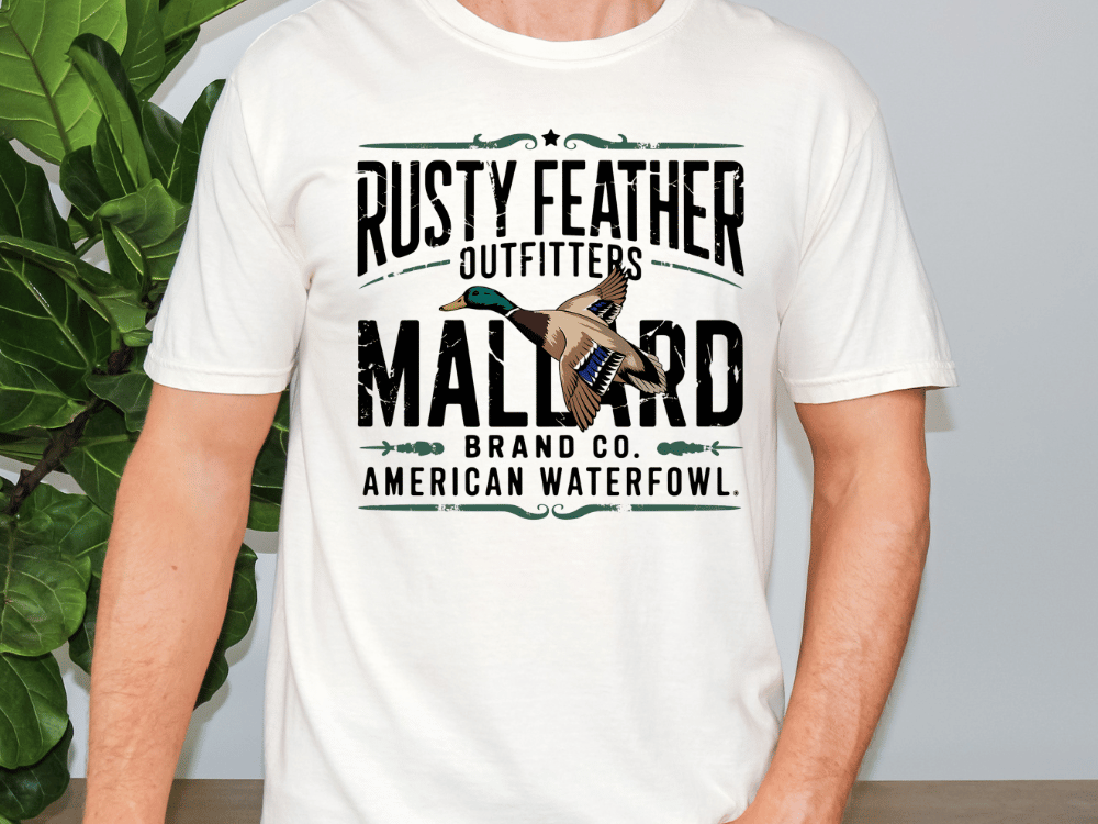 A white t-shirt with a graphic design featuring a bird, specifically a mallard, and text that reads "RUSTY FEATHER OUTFITTERS MALARD BRAND CO. AMERICAN WATERFOWL".