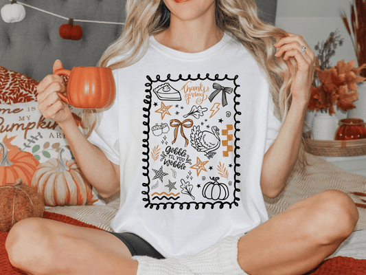 A woman sitting on a bed, holding a mug and wearing a white t-shirt with a graphic design featuring various fall-themed elements such as pumpkins, leaves, and a turkey.