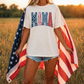 A woman stands in a field, holding an American flag and wearing a white t-shirt with the word "Mama" printed on it.