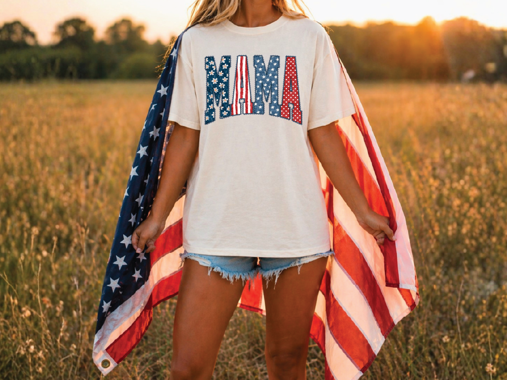 A woman stands in a field, holding an American flag and wearing a white t-shirt with the word "Mama" printed on it.