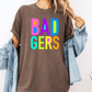A person wearing a brown t-shirt with the text "BAD GERS" in colorful letters, paired with a denim jacket.