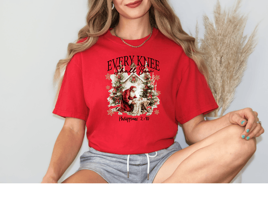 A woman wearing a red t-shirt with a Christmas-themed graphic and text, sitting with her legs crossed.