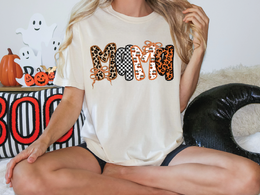 A woman wearing a white t-shirt with the word "MOM" printed on it, sitting cross-legged on the floor. There are Halloween decorations and ghost figures in the background.