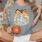 A woman wearing a gray t-shirt with a pumpkin graphic and holding a mug is sitting on a bed with autumn-themed pillows.