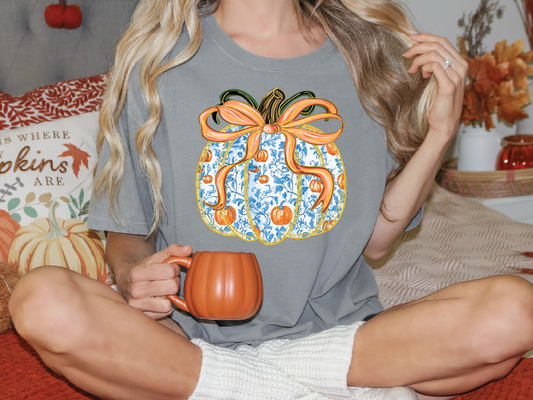 A woman wearing a gray t-shirt with a pumpkin graphic and holding a mug is sitting on a bed with autumn-themed pillows.