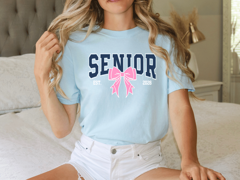 A woman wearing a light blue t-shirt with a pink bow and the text "SENIOR" printed on it, sitting on a bed.