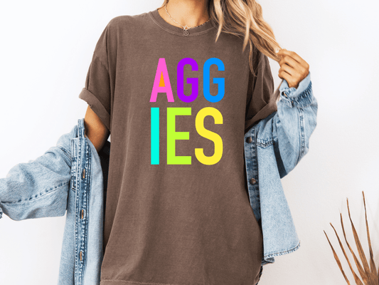 A person wearing a brown t-shirt with the text "AGGIES" in colorful letters, paired with a denim jacket, stands against a white background.