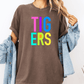 A person wearing a brown t-shirt with the word "TIGERS" in colorful letters, paired with a denim jacket, stands against a white background.