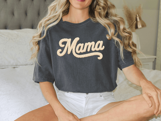 A woman with long blonde hair wearing a black t-shirt with the word "Mama" printed on it, sitting on a bed.