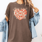 A woman wearing a brown t-shirt with a heart design and text, along with a denim jacket, stands against a white background.