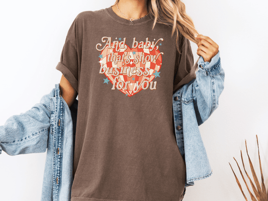 A woman wearing a brown t-shirt with a heart design and text, along with a denim jacket, stands against a white background.
