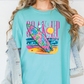 A woman wearing a turquoise t-shirt with a colorful graphic of a surfboard and beach scene.