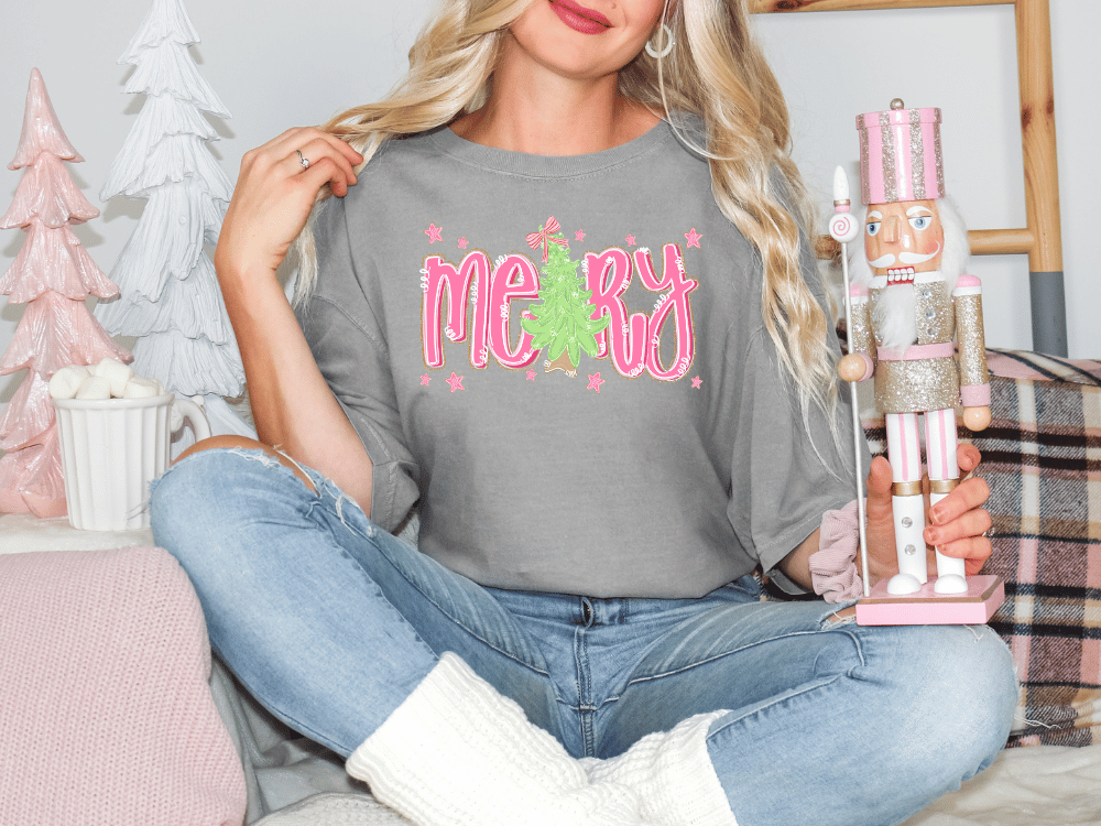 A woman sitting on a couch holding a nutcracker and wearing a gray t-shirt with a Christmas tree design.