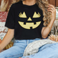 A woman wearing a black t-shirt with a pumpkin jack-o-lantern design is sitting on a couch surrounded by fall decorations.