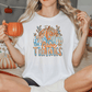 A woman sitting on a bed, holding a mug and wearing a white t-shirt with a pumpkin graphic and the text "Give Thanks".