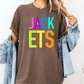 A person wearing a brown t-shirt with the text "JACK ETS" in colorful letters, along with a denim jacket, stands against a white background.