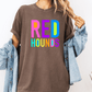 A person wearing a brown t-shirt with the text "RED HOUNDS" in colorful letters, along with a denim jacket, stands against a white background.