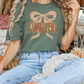 A person wearing a green t-shirt with a leopard print bow and the word "THANKFUL" is sitting on a surface surrounded by fall decorations, including pumpkins and a lantern.