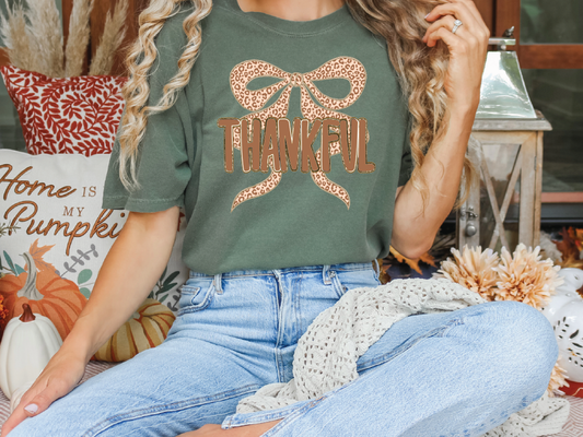A person wearing a green t-shirt with a leopard print bow and the word "THANKFUL" is sitting on a surface surrounded by fall decorations, including pumpkins and a lantern.
