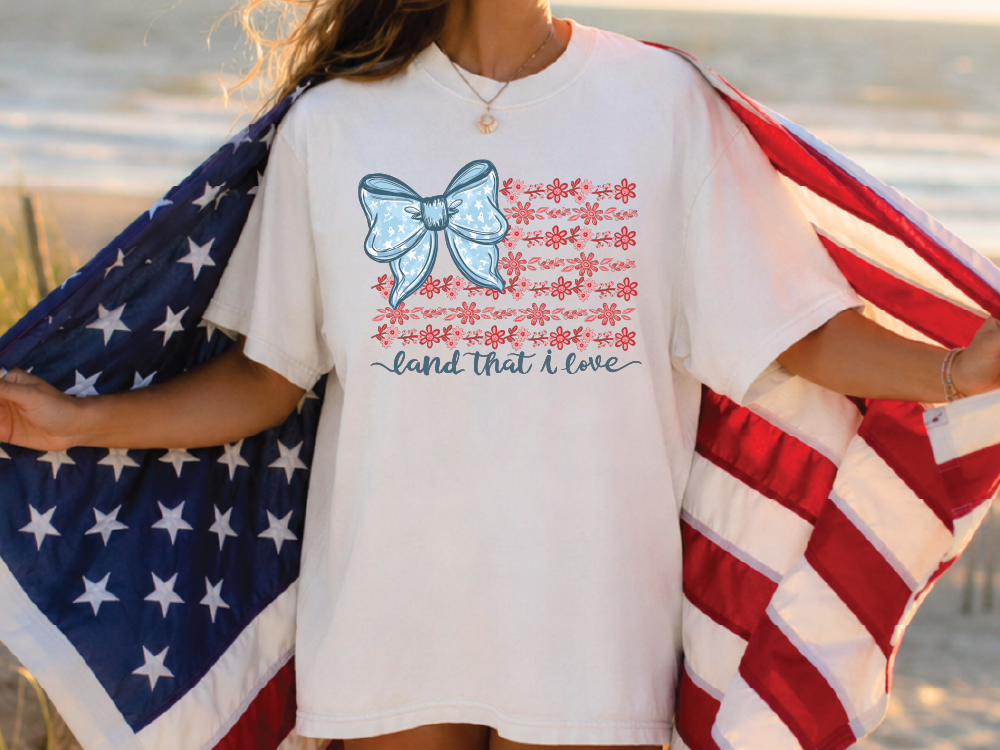 A person wearing a white t-shirt with a graphic design of an American flag and the text "land that I love" printed on it, holding the flag in their arms.