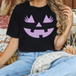 A woman wearing a black t-shirt with a purple jack-o-lantern graphic is sitting on a couch surrounded by fall decorations.