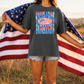 A person standing in a field holding an American flag, wearing a gray t-shirt with a graphic design.