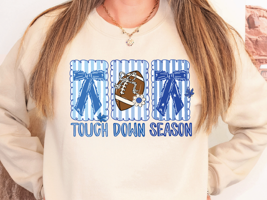 A woman wearing a white sweatshirt with a graphic design featuring a football, blue bows, and the text "TOUGH DOWN SEASON".