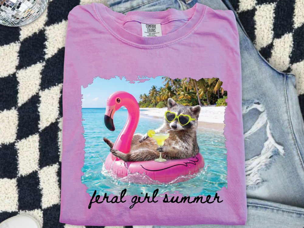 A pink t-shirt with a graphic design depicting a raccoon wearing sunglasses and floating on a pink flamingo in a tropical beach setting.