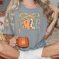 A woman wearing a gray t-shirt with the word "FALL" printed on it, sitting on a bed with a pumpkin-themed pillow and holding a mug.