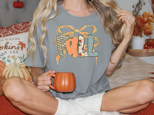 A woman wearing a gray t-shirt with the word "FALL" printed on it, sitting on a bed with a pumpkin-themed pillow and holding a mug.
