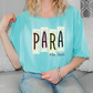 A woman wearing a turquoise t-shirt with the word "PARA" printed on it, sitting on a couch.