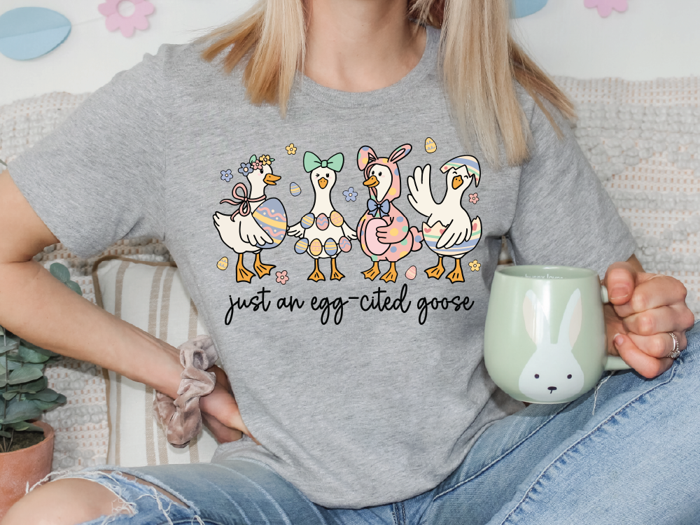 A woman wearing a gray t-shirt with a graphic of four cartoon ducks and the text "Just an egg-cited goose" is sitting on a couch, holding a green mug.