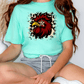 A person wearing a turquoise t-shirt with a graphic of a rooster and a leopard print design, sitting on the floor.