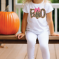 A young girl wearing a white t-shirt with a leopard print design and the word "BOO" is sitting on a wooden bench next to a large orange pumpkin.