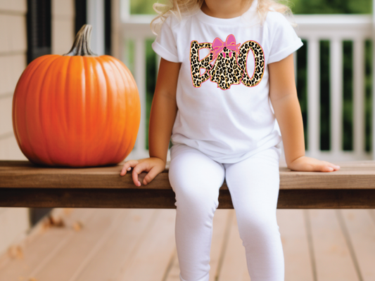 A young girl wearing a white t-shirt with a leopard print design and the word "BOO" is sitting on a wooden bench next to a large orange pumpkin.