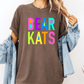 A person wearing a brown t-shirt with the text "BEAR KATS" in colorful letters, along with a denim jacket, stands against a white background.