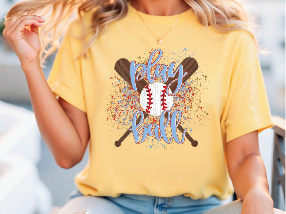 A woman wearing a yellow t-shirt with a graphic design of a baseball bat and ball, along with the text "Play ball" printed on it.