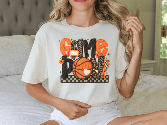 A woman wearing a white t-shirt with a basketball graphic and the text "Game Day" is sitting on a bed.