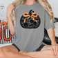 A woman is sitting cross-legged on a couch, wearing a gray t-shirt with a black pumpkin design and an orange ribbon bow.