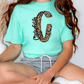 A person wearing a turquoise t-shirt with a leopard print design and gray shorts is sitting on the floor.